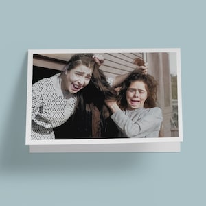 May include: Two women with long dark hair are pulling each other's hair. They are both wearing white and black checkered dresses. The woman on the left is smiling and the woman on the right is crying.