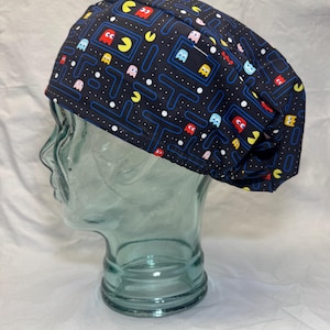May include: A dark blue surgical scrub cap featuring a colorful Pac-Man video game pattern. The cap has a repeating design of Pac-Man characters and mazes in yellow, red, blue, and pink, with white dots on a dark blue background.