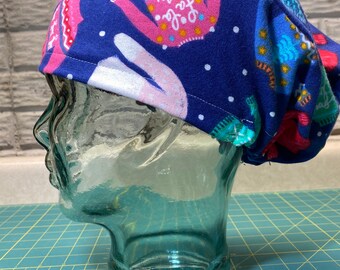 May include: Blue scrub hat with a white and pink holiday sweater pattern. The hat is on a glass head form.