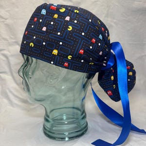 Puede incluir: Un gorro quirúrgico azul marino con un diseño de videojuego Pac-Man, con fantasmas coloridos y personajes de Pac-Man. El gorro tiene un coletero a juego y está atado con una cinta de satén azul brillante.