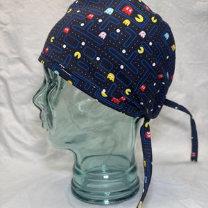 Puede incluir: Un gorro quirúrgico azul marino con un colorido diseño del videojuego Pac-Man. El gorro, con cierre de lazo, se muestra en una cabeza de maniquí transparente. El diseño incluye personajes de Pac-Man y elementos de laberinto.
