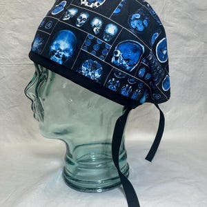 May include: A black surgical scrub cap with a repeating pattern of blue skull imagery. The cap has a black band and ties. The skull images are in various sizes and orientations, creating a medical or anatomical theme.