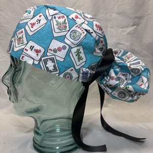 Peut inclure: Un bonnet de gommage à motifs bleus et blancs avec un motif de tuiles de Mahjong. Le bonnet a un dos froncé et un lien en ruban noir. Les tuiles de Mahjong sont blanches avec des détails rouges, verts et bleus.