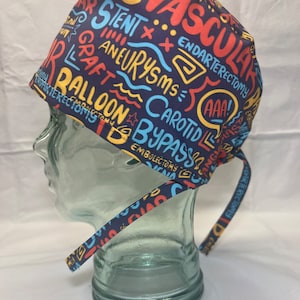 May include: Navy blue surgical scrub cap with colorful text including "Vascular", "Stent", "Aneurysms", and "Balloon". The cap has a tie closure and is designed for medical professionals.