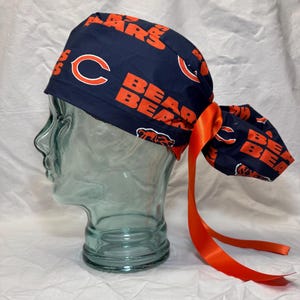 May include: A navy blue scrub cap featuring an all-over print of the Chicago Bears logo and the word BEARS in orange lettering. The cap includes a matching orange satin ribbon tie at the back to secure the fit, with a gathered fabric pouch.