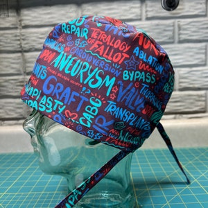 May include: A dark blue and red surgical scrub hat with a colorful print of medical terms, including "repair", "tetralogy of fallot", "aneurysm", "graft", "valve", and "transplant".