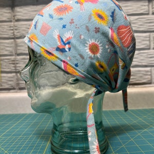 Pode incluir: Um gorro cirúrgico azul com estampa floral e de pássaros coloridos. O gorro tem uma gravata que pode ser amarrada na parte de trás do pescoço.