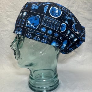 May include: A blue and black surgical scrub cap featuring a repeating pattern of medical imaging scans. The images are in shades of blue and white, set against a black background. The cap is designed to fit comfortably.