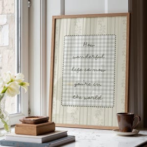Puede incluir: Un estampado enmarcado con el texto "How wonderful life is now you're in the world." El estampado tiene un marco de madera marrón claro y un patrón de cuadros grises y blancos. El fondo tiene un diseño floral.