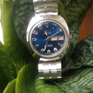 Puede incluir: Un reloj Oris plateado con una esfera azul y agujas plateadas. El reloj tiene una ventana de fecha y una ventana de día de la semana. El reloj está en una banda de metal plateado.
