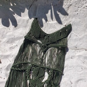 Puede incluir: Un top de macram&eacute; verde oscuro con un dise&ntilde;o geom&eacute;trico y detalles de flecos. El top est&aacute; colocado sobre una superficie de arena blanca.