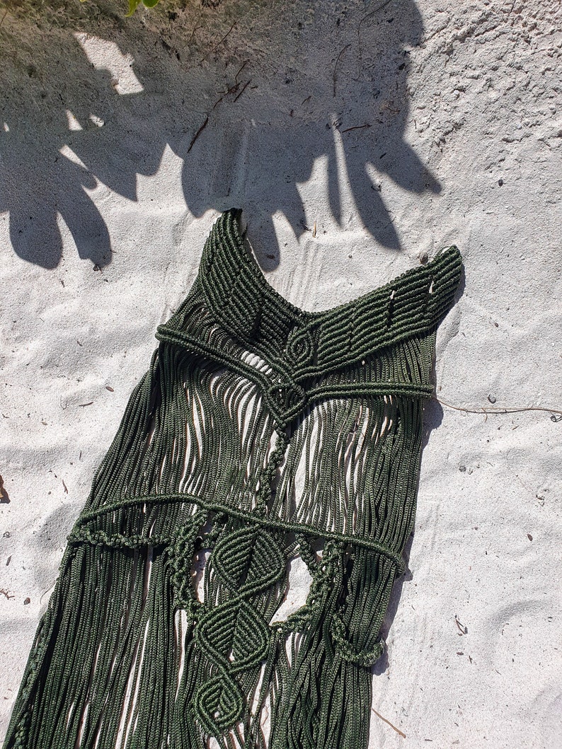 Puede incluir: Un top de macram&eacute; verde oscuro con un dise&ntilde;o geom&eacute;trico y detalles de flecos. El top est&aacute; colocado sobre una superficie de arena blanca.