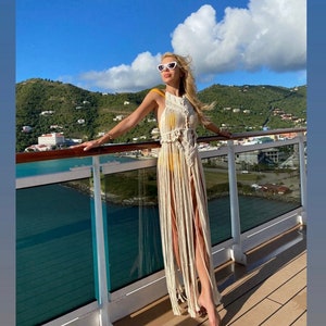 Puede incluir: Una mujer con un vestido largo de crochet blanco con detalles de flecos, de pie en la cubierta de un crucero con vistas a una isla tropical.