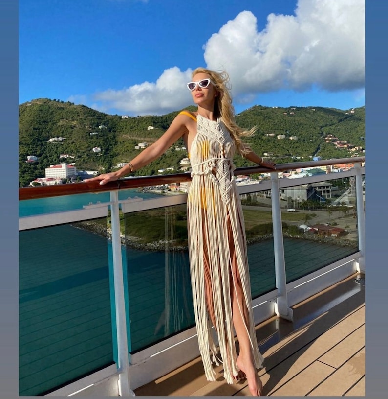 Puede incluir: Una mujer con un vestido largo de crochet blanco con detalles de flecos, de pie en la cubierta de un crucero con vistas a una isla tropical.