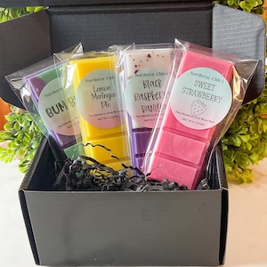 May include: A black box filled with four wax melts. The wax melts are wrapped in clear plastic and have labels with the names "Lemon Meringue Pie", "Black Raspberry Vanilla", "Sweet Strawberry", and "Bum Bum".