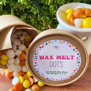 May include: A container of wax melt dots with a colorful label that reads "WAX MELT DOTS". The dots are in various colors, including white, yellow, and orange. A small bowl of wax melts is in the background.