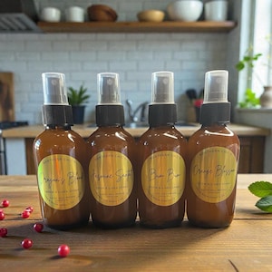 May include: Four brown glass spray bottles with black caps and gold labels, labeled "Dragon's Blood", "Jasmine Secret", "Bum Bum", and "Orange Blossom". The bottles are arranged on a wooden surface with scattered red berries.