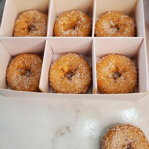 May include: A white box with six small, round donuts with sugar sprinkles. The donuts are arranged in two rows of three. The box is open and the donuts are visible.