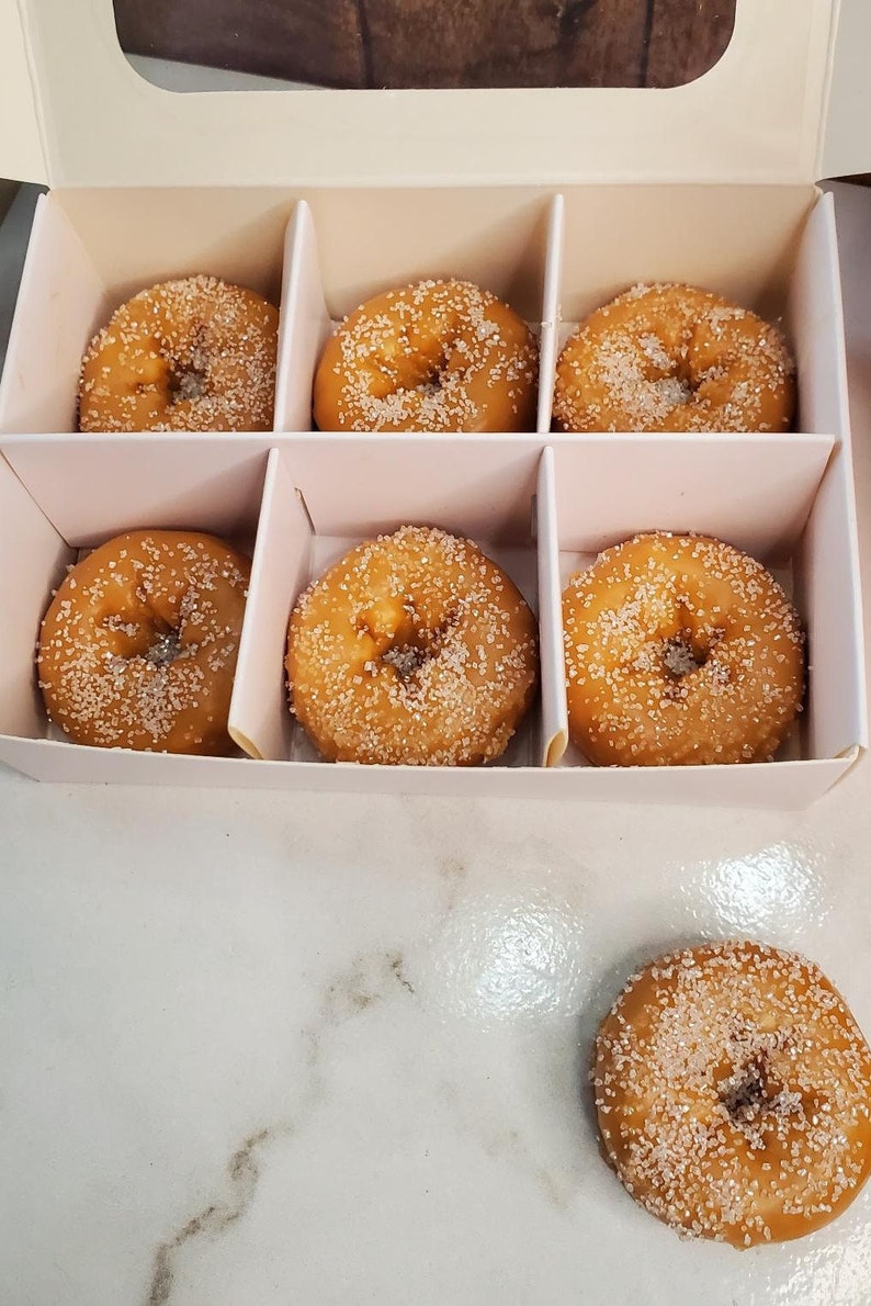 May include: A white box with six small, round donuts with sugar sprinkles. The donuts are arranged in two rows of three. The box is open and the donuts are visible.