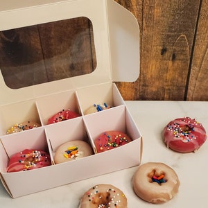 May include: A white box with a clear window, containing six mini donuts with pink and white frosting and colorful sprinkles. Three additional donuts are outside the box, two with white frosting and one with pink frosting.