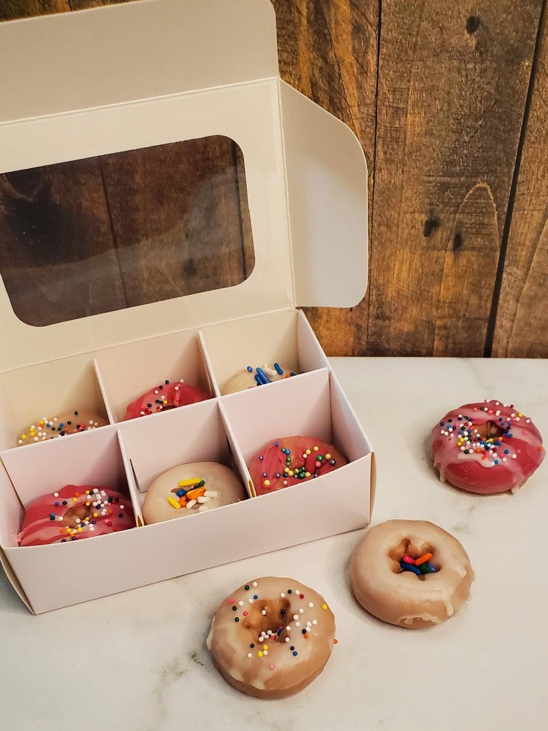 May include: A white box with a clear window, containing six mini donuts with pink and white frosting and colorful sprinkles. Three additional donuts are outside the box, two with white frosting and one with pink frosting.