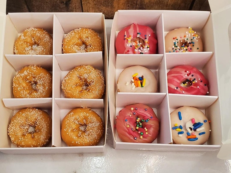 May include: A white box with 10 donuts in it. The donuts are arranged in two rows of five. The donuts are glazed, sprinkled, and have different colored icings.
