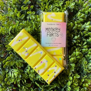 May include: A yellow wax melt bar with white swirls and glitter. The bar is packaged in a clear plastic wrapper with a label that reads "Northern Chic's Monkey Farts Handmade in Ohio Wax Melt Net Wt 1.75 oz".