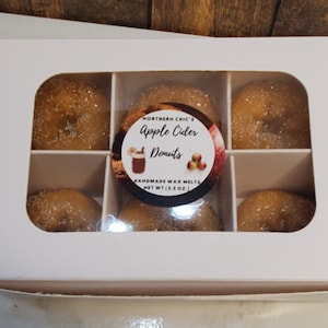 May include: A white box with six individual compartments containing small, round, sugared donuts. The box has a label that reads "Northern Chic's Apple Cider Donuts Handmade Wax Melts Net Wt (3.5 oz)"