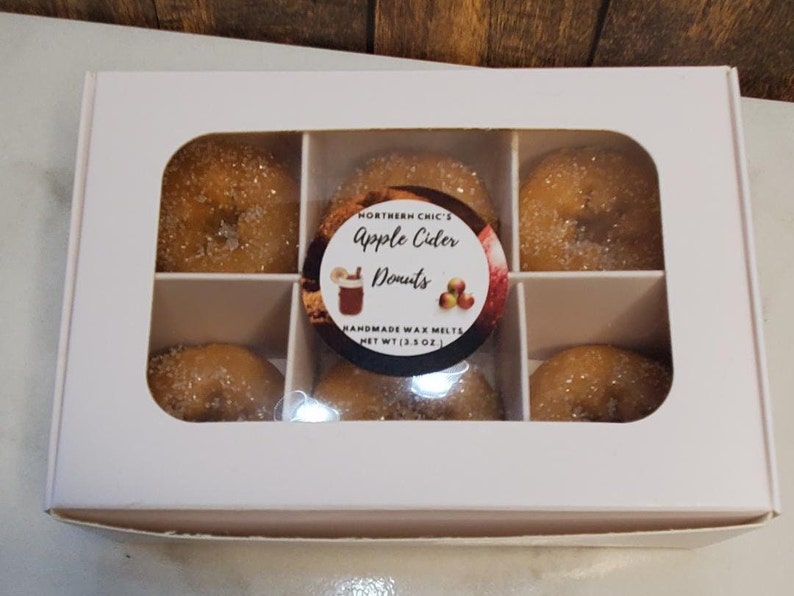 May include: A white box with six individual compartments containing small, round, sugared donuts. The box has a label that reads "Northern Chic's Apple Cider Donuts Handmade Wax Melts Net Wt (3.5 oz)"