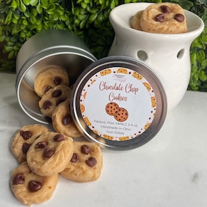 May include: A silver tin filled with chocolate chip cookie wax melts. The tin has a label that reads "Chocolate Chip Cookies" and "Parasoy Wax Melts | 3-4 oz. Handmade in Ohio. Non-Edible."