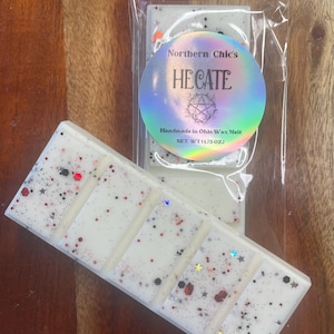 May include: Two rectangular white wax melt bars with five segments each, speckled with red, black, and silver glitter. A circular holographic label on the clear plastic packaging reads Northern Chic's HECATE Handmade in Ohio Wax Melt NET WT 1.75 OZ.