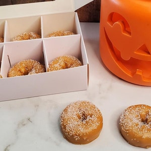 May include: A white box with six small, round, sugar-coated donuts inside. Two additional donuts are on a white surface next to the box. A large orange pumpkin-shaped container is in the background.