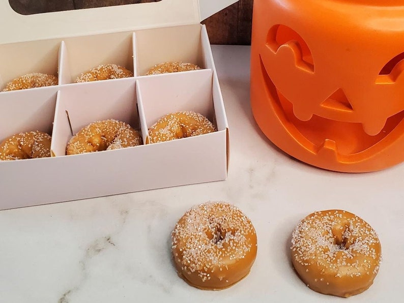 May include: A white box with six small, round, sugar-coated donuts inside. Two additional donuts are on a white surface next to the box. A large orange pumpkin-shaped container is in the background.