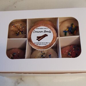 May include: A white box with six clear windows showing six small wax melts shaped like donuts. The wax melts are topped with colorful sprinkles. The label on the box reads "Northern Chic's Cinnamon Donuts Handmade Wax Melts Net Wt (1.3 oz)"