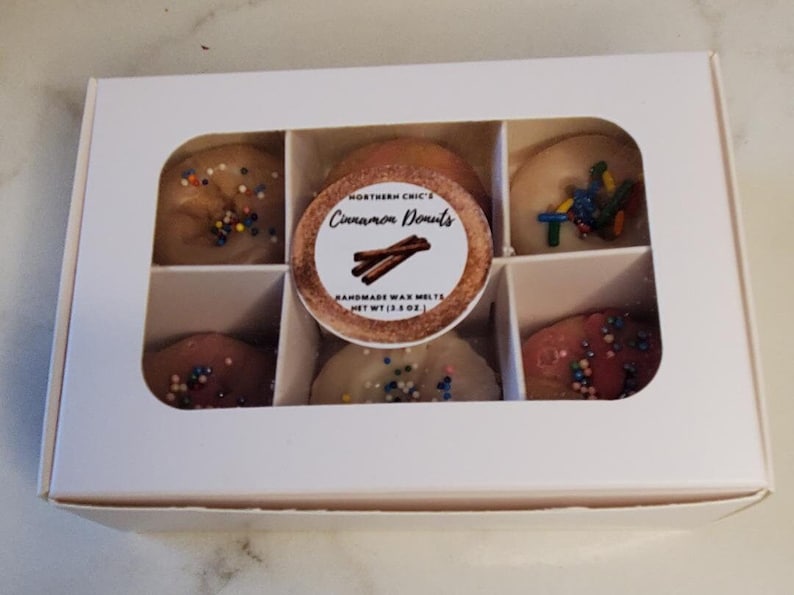 May include: A white box with six clear windows showing six small wax melts shaped like donuts. The wax melts are topped with colorful sprinkles. The label on the box reads "Northern Chic's Cinnamon Donuts Handmade Wax Melts Net Wt (1.3 oz)"