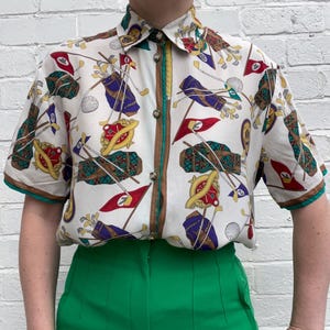 May include: A short-sleeved button-down shirt with a golf-themed pattern, featuring golf bags, flags, and golf balls on a cream background. The shirt has a green trim and is paired with bright green pants.