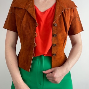 Könnte beinhalten: Kurzärmelige braune Wildlederjacke mit Kragen und Knopfverschlüssen. Die Jacke wird mit einem leuchtend orangefarbenen Oberteil und einer grünen Hose kombiniert. Die Jacke hat dekorative Ausschnitte an den Ärmeln.