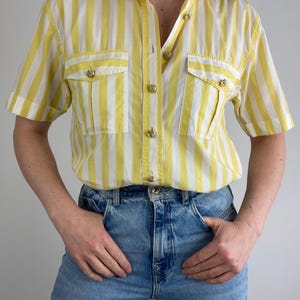 May include: A striped shirt in yellow and white, with short sleeves and a button-down design. It features two front pockets and gold-toned buttons. The shirt is worn with light blue denim jeans.