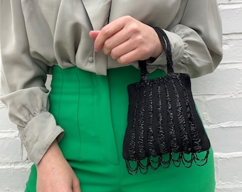 Op de afbeelding: Een grijs blouse met een gerimpelde kraag en lange mouwen, gecombineerd met een groene broek. Een klein zwart kralen tasje met een franje detail hangt aan de hand van de persoon.