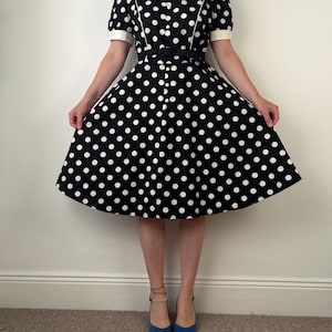 May include: A black knee-length dress featuring a white polka dot pattern, short sleeves with white cuffs, a round neckline, a front button placket, and a black belt at the waist. The skirt has a flared silhouette with white piping details.