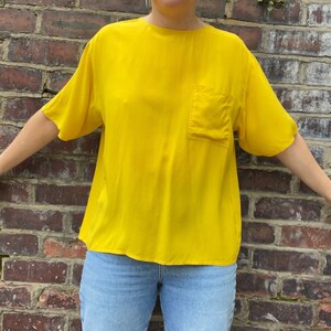 May include: A bright yellow short-sleeved blouse with a single pocket on the left side. The blouse is made of a silky material and has a relaxed fit.