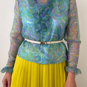 May include: A long-sleeve blouse featuring a sheer fabric with a swirling blue, green, and purple pattern, a ruffled collar, and button-down front. The blouse is cinched at the waist with a thin, textured white belt and paired with a bright yellow pleated skirt.