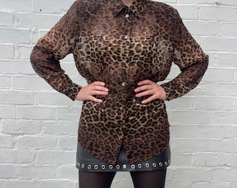 Op de afbeelding: Een doorschijnende blouse met luipaardprint in bruin en zwart, met een knoopsluiting en lange mouwen. De blouse heeft een klassieke kraag en manchetten met knopen. Gedragen met een zwarte rok en zwarte panty's.
