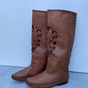 Puede incluir: Botas de cuero marrón hasta la rodilla con diseños recortados y un detalle tejido en la parte posterior del eje.