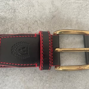 May include: A black leather belt with a gold buckle and red stitching. The belt has a logo with a shield and a laurel wreath.