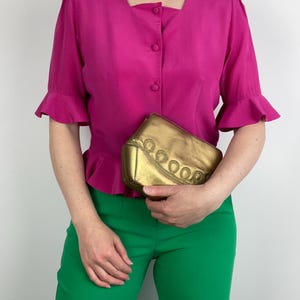 May include: A magenta blouse with a square neckline and button closure, worn with green trousers. A gold clutch with an ornate design is held in front. The blouse has short sleeves with ruffled cuffs.