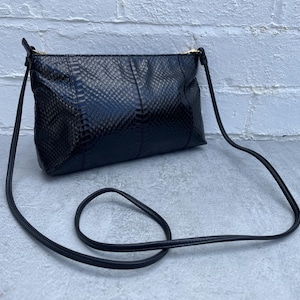 May include: Black crossbody bag with a textured, snake-like pattern. The bag has a long, thin strap and a gold-coloured zip. The bag is rectangular and appears to be made of leather or a similar material.