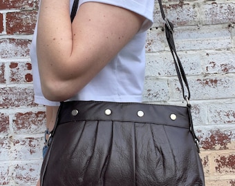 May include: A brown leather shoulder bag with a pleated front and silver studs. The bag has a long adjustable strap.