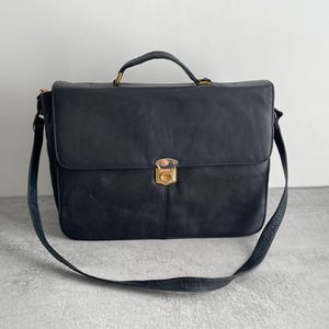 May include: A black leather briefcase with a top handle and a detachable shoulder strap. The bag features a gold-coloured clasp and hardware. The briefcase is rectangular and appears to be made of a soft, supple leather.