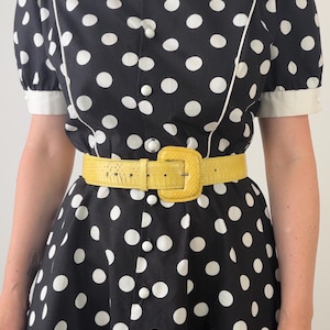 May include: A black dress with white polka dots, short puff sleeves, and white trim. The dress features a front button closure with round white buttons. A bright yellow textured belt with a matching yellow buckle is worn around the waist.